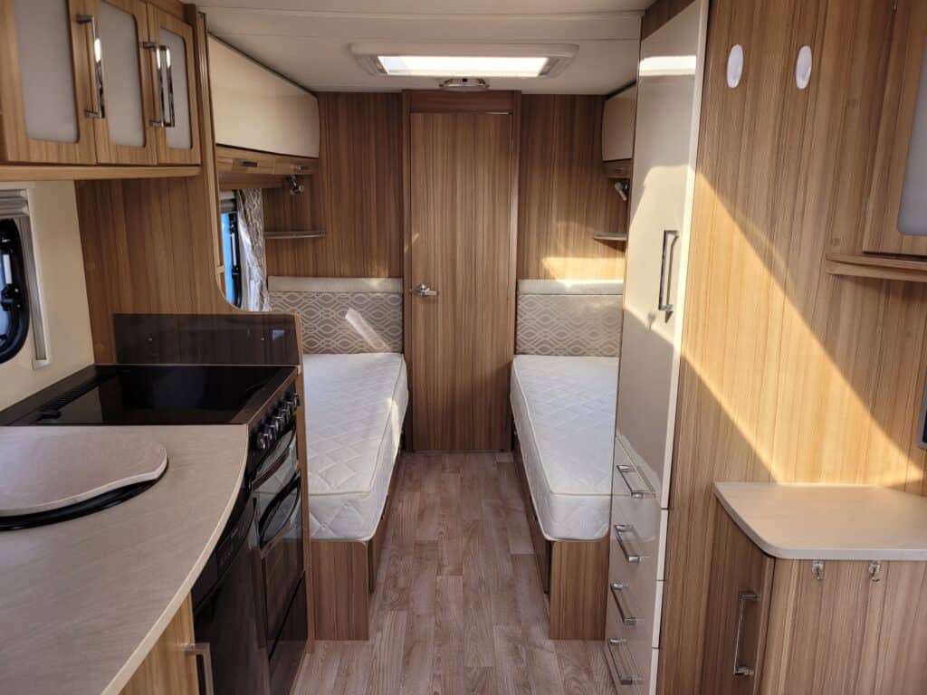 Interior view of a campervan with twin beds, wooden storage units, and a small kitchen area.